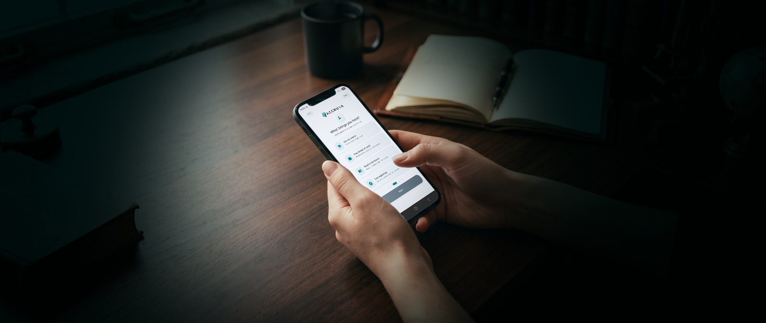 Hands holding a phone showing the Accruva onboarding screen, with a coffee mug and open leather notebook visible in soft focus on a dark wooden desk under warm window light