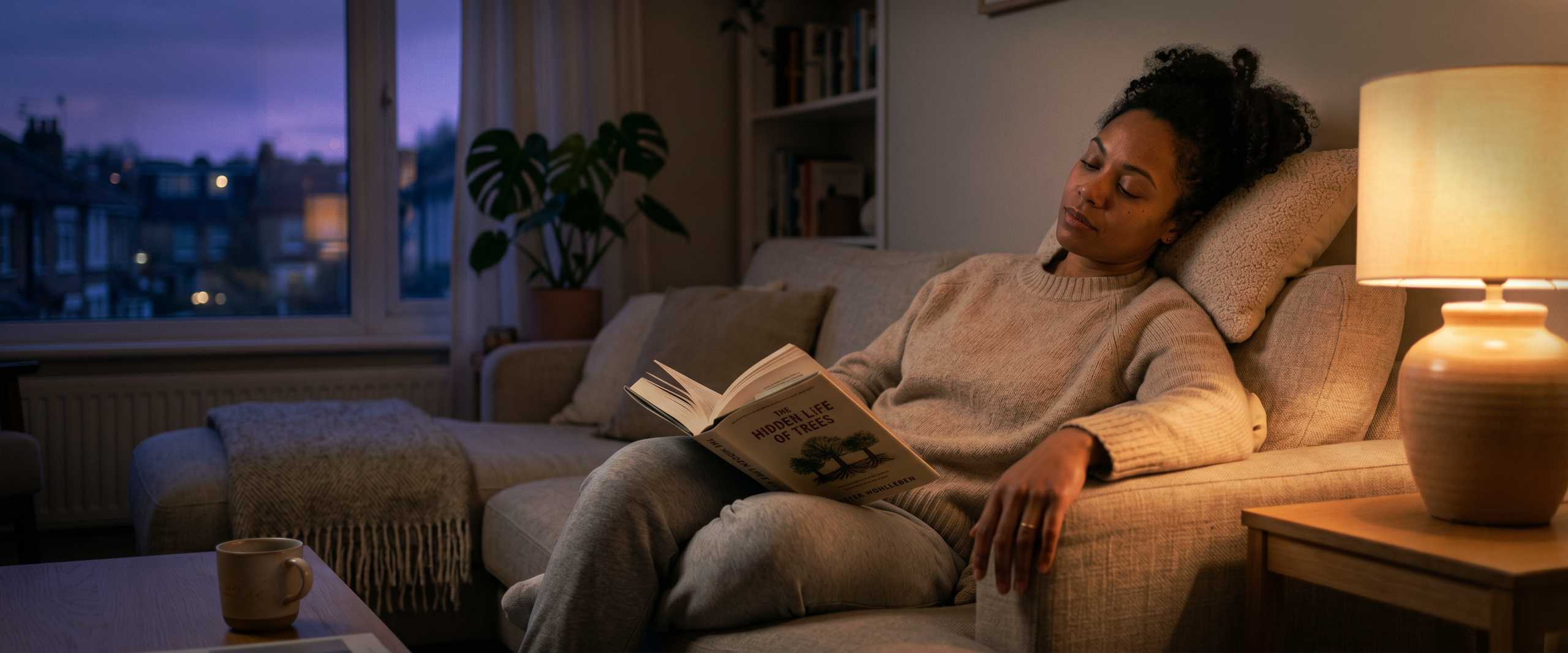 An adult on a sofa in lamp-lit evening light, a book open on their lap, head resting back against the cushion in late-day cognitive fatigue